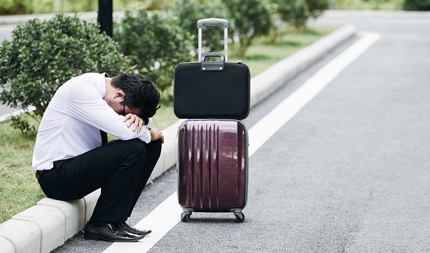 A person sitting on a curb with his head down, with luggage on his side.