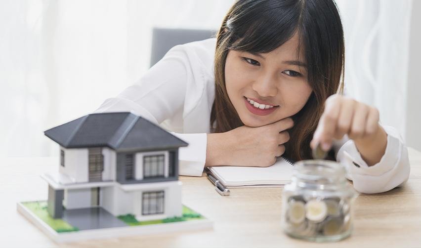 A young woman smiles while placing coins into a jar, with a model house on the table, symbolizing the use of home equity loans (HELOC) to fund EB5 investments.