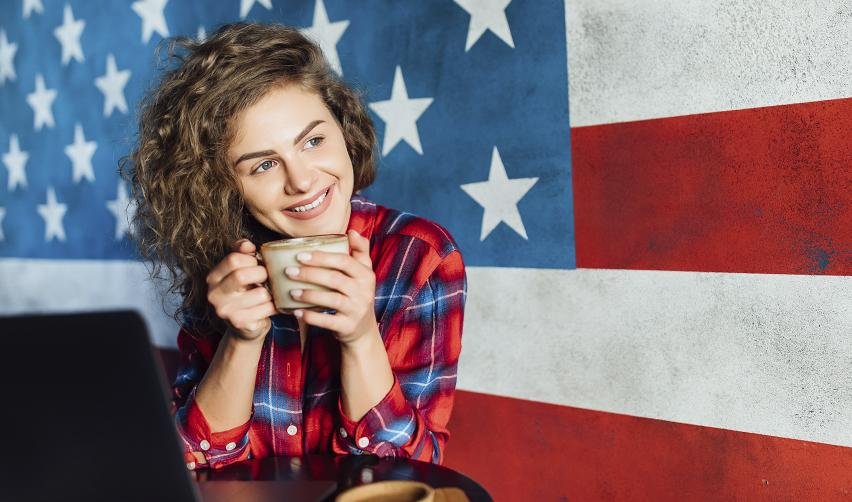 A smiling young woman in front of a large American flag mural, representing the freedom and comfort of living in the U.S. while awaiting EB5 Green Card approval.