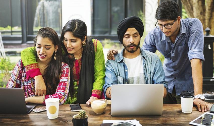 Group of young Indian professionals collaborating on laptops in an outdoor setting, discussing immigration options through a sponsoring employer that USCIS approves to obtain permanent residence.
