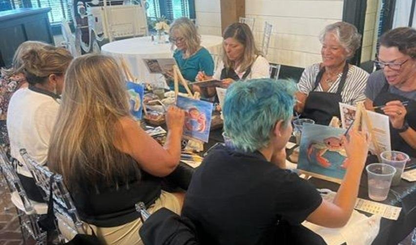 Group of Bay Creek residents smiling while painting coastal scenes at a guided art class, part of the community’s creative lifestyle programs.